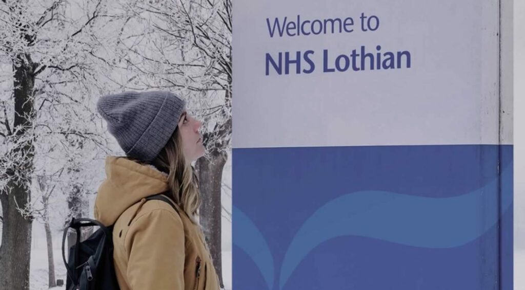 A young woman in the snow in front of a sign reading Welcome to NHS Lothian