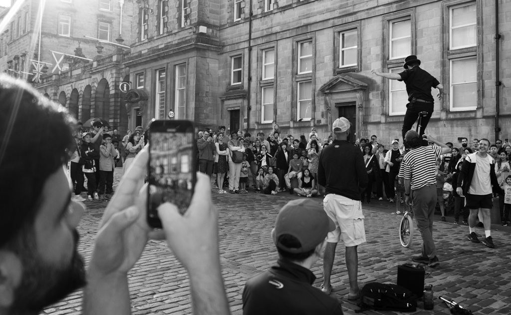 A crowd watched a street performer in Edinburgh. A man in the foreground is capturing the scene using his mobile phone.
